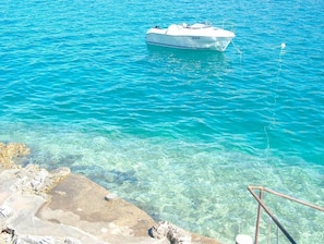 A boat anchored in clear turquoise water with a rocky shoreline in the foreground. - Guest House Blanka (Okrug)