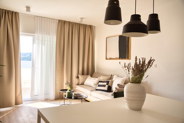 A modern living room with a sofa, a coffee table, and a vase of plants. There is a large window with curtains, and a mirror on the wall. - Guest House Blanka (Okrug)