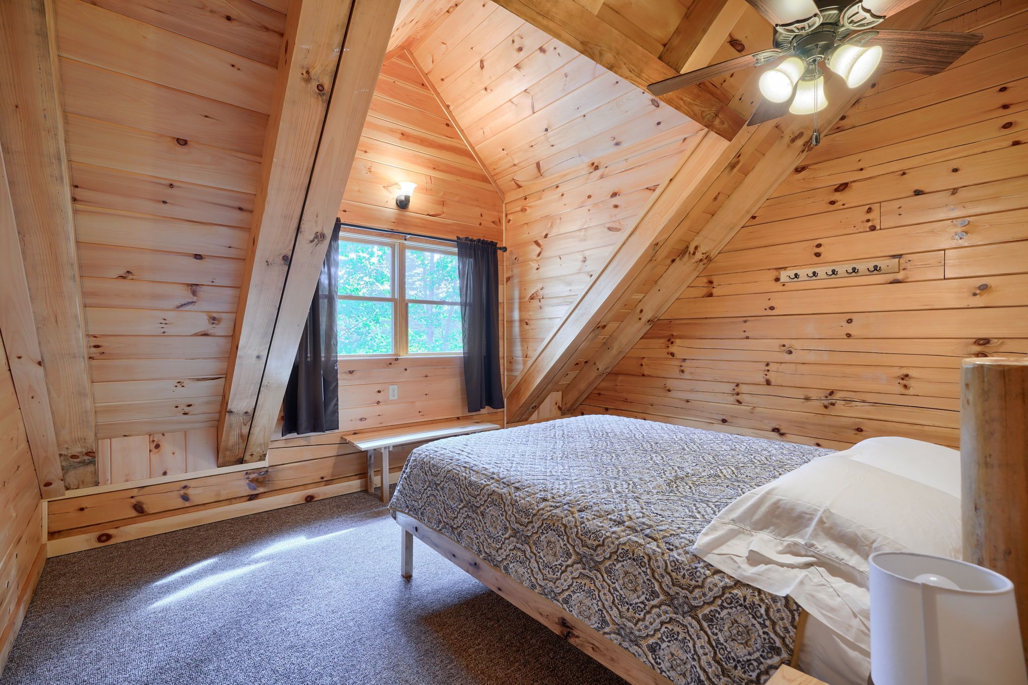 A wooden cabin bedroom with a bed, a bench, a window with curtains, and a ceiling fan.