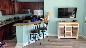 A kitchen with dark wood cabinets, a granite countertop, a stainless steel refrigerator, a microwave, and a stove. There are two bar stools and a small wooden cabinet with drawers. - ~Carolina Tides~ Ocean View, Pier, Pool, Steps to the Beach and Restaurants! (Oak Island)