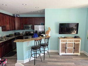 A kitchen with dark wood cabinets, a granite countertop, a stainless steel refrigerator, a microwave, and a stove. There are two bar stools and a small wooden cabinet with drawers.