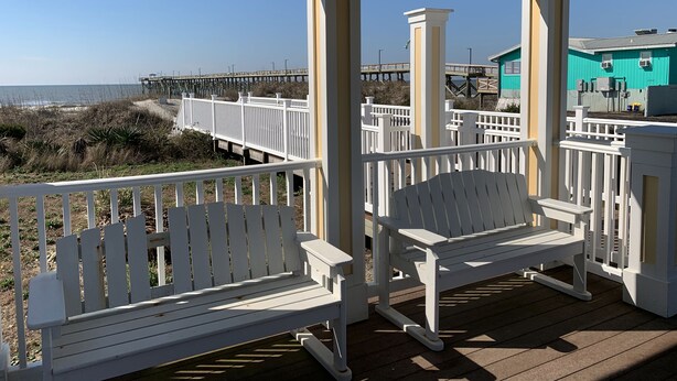 A covered deck with white railings and benches overlooking a beach with a wooden pier and a turquoise building in the background. - ~Carolina Tides~ Ocean View, Pier, Pool, Steps to the Beach and Restaurants! (Oak Island)
