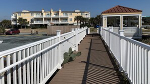 A wooden boardwalk with white railings, leading to a building with balconies and a covered area. - ~Carolina Tides~ Ocean View, Pier, Pool, Steps to the Beach and Restaurants! (Oak Island)