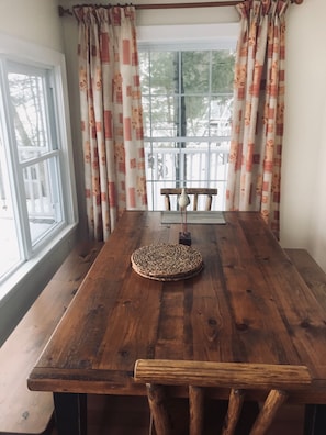 Una mesa de comedor de madera con un camino de mesa tejido, una silla de madera y una ventana con cortinas florales.