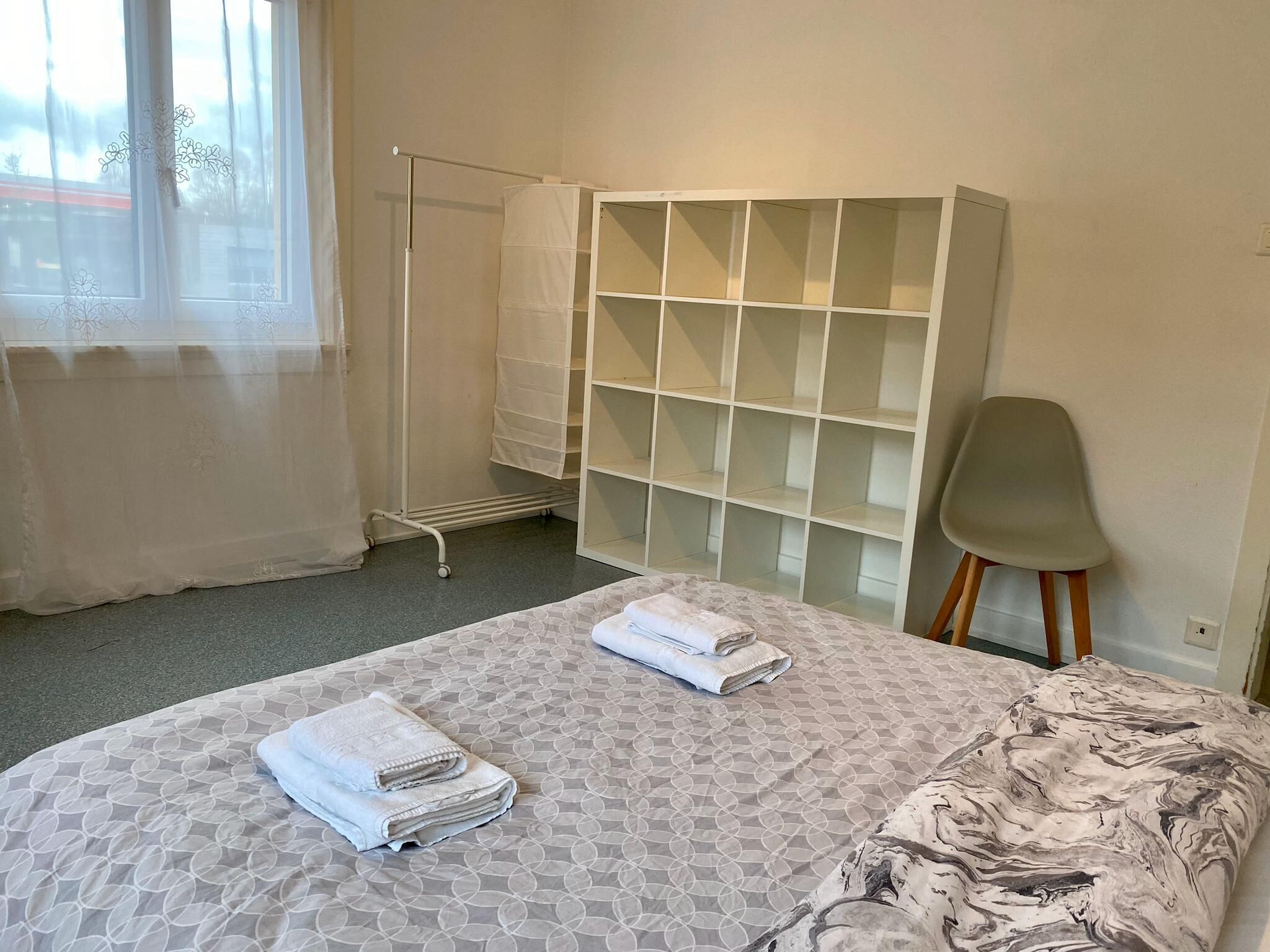 A hotel room with a bed, a wardrobe, a chair, and a window with sheer curtains.