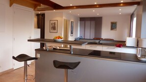 Eine moderne Küche mit einer zentralen Kochinsel, zwei Barhocker und einem Essbereich mit Blick auf das Wohnzimmer.