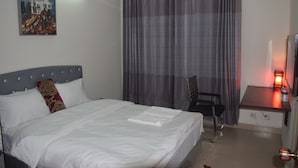 A hotel room with a bed, a desk with a lamp, a chair, and a painting on the wall. - Fairmont Guest House (Dhaka)