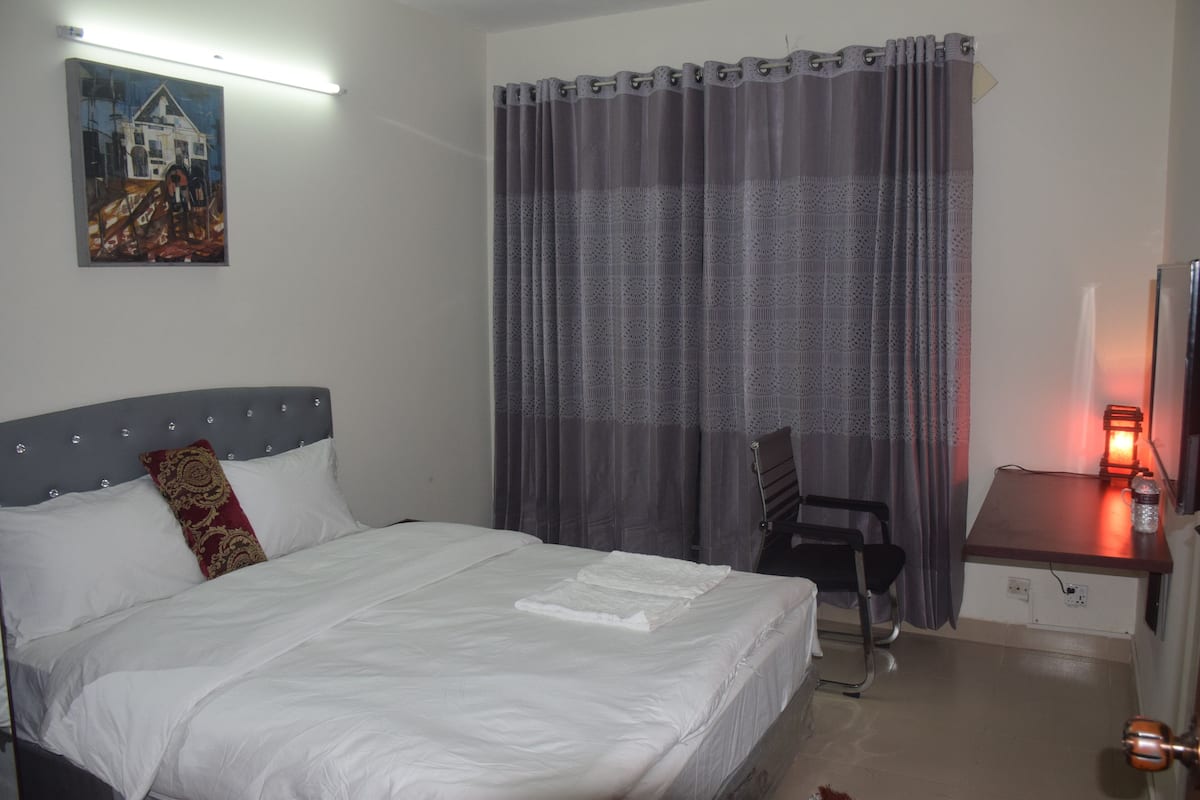 Ett hotellrum med en säng, ett skrivbord med en lampa, en stol och en tavla på väggen.