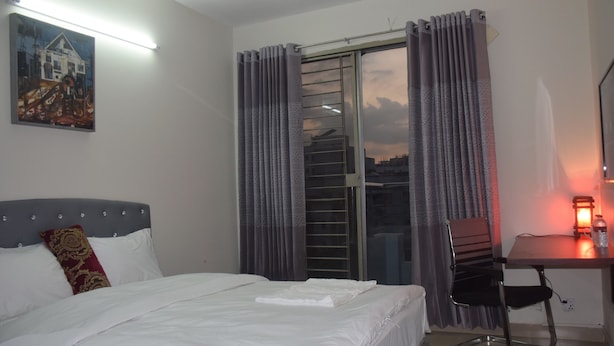 A hotel room with a bed, a desk, a chair, a window with curtains, and a painting on the wall. - Fairmont Guest House (Dhaka)