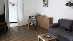 A living room with a grey sofa, wooden cabinets, a small table, and a standing chair with a patterned cover. - Near Royan, 96m2 house with 500m2 enclosed garden - 3 beaches within 600m (Vaux-sur-Mer)