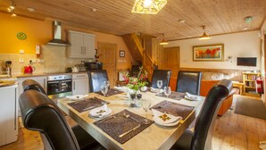 A dining area with a wooden table set for six, surrounded by black leather chairs. A kitchen with white cabinets and appliances is visible in the background.