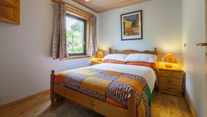 A bedroom with a wooden bed, a window with curtains, a nightstand with a lamp, and a painting on the wall.