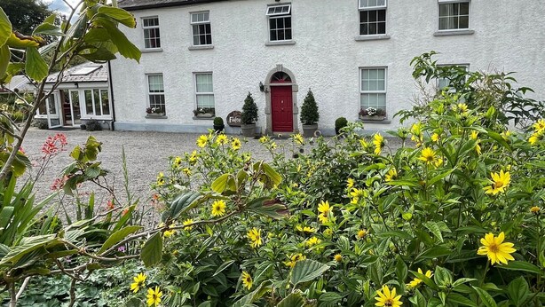 A white building with a red door, multiple windows, and a garden with yellow flowers in the foreground.