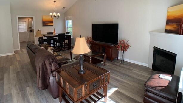 A spacious living room with a high ceiling fan, a fireplace, a flat-screen TV, and a dining area with a chandelier. - Parris Island Marine Corps Graduation PCS Orders Travel Nurses Beaufort Home (Beaufort)