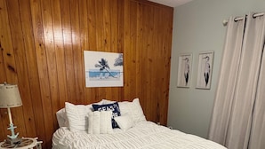 Quarto com parede revestida em madeira, cama com roupa de cama branca, ventoinha no teto e uma pequena mesa com abajur.