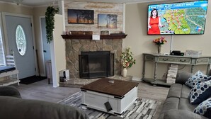 Uma sala de estar com lareira de pedra, televisão na parede exibindo a previsão do tempo e sofá com almofadas estampadas.