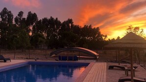 Une piscine avec une terrasse en bois, entourée d’arbres, et un coucher de soleil en arrière-plan.