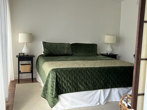 Una cama bien hecha con colcha verde, dos mesitas de noche con lámparas y una pequeña cesta de mimbre sobre una de ellas.