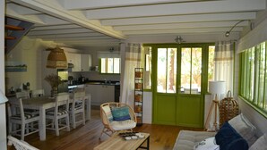 Un vaste espace de vie comprenant une table à manger en bois, un canapé et un fauteuil en osier. On aperçoit une cuisine au fond, avec une porte verte donnant sur l’extérieur.