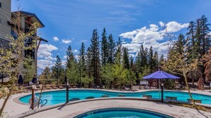 A swimming pool with a hot tub, surrounded by lounge chairs and umbrellas, set against a backdrop of trees and a clear sky. - Quiet Village Monache Retreat | Balcony Views, Fireplace, Walk to Gondola (Mammoth Lakes)