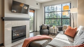A modern living room with a fireplace, comfortable seating, and a large window offering a view of trees. - Quiet Village Monache Retreat | Balcony Views, Fireplace, Walk to Gondola (Mammoth Lakes)