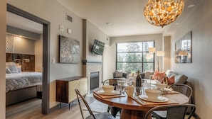 A modern living room with a dining area, a fireplace, and a large window offering a view of the outdoors. - Quiet Village Monache Retreat | Balcony Views, Fireplace, Walk to Gondola (Mammoth Lakes)