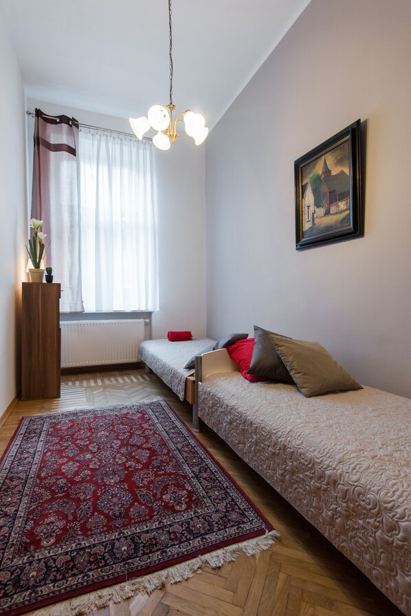 Un dormitorio con dos camas individuales, una alfombra roja estampada, una mesita de madera con una planta, un radiador y una ventana con cortinas translúcidas.