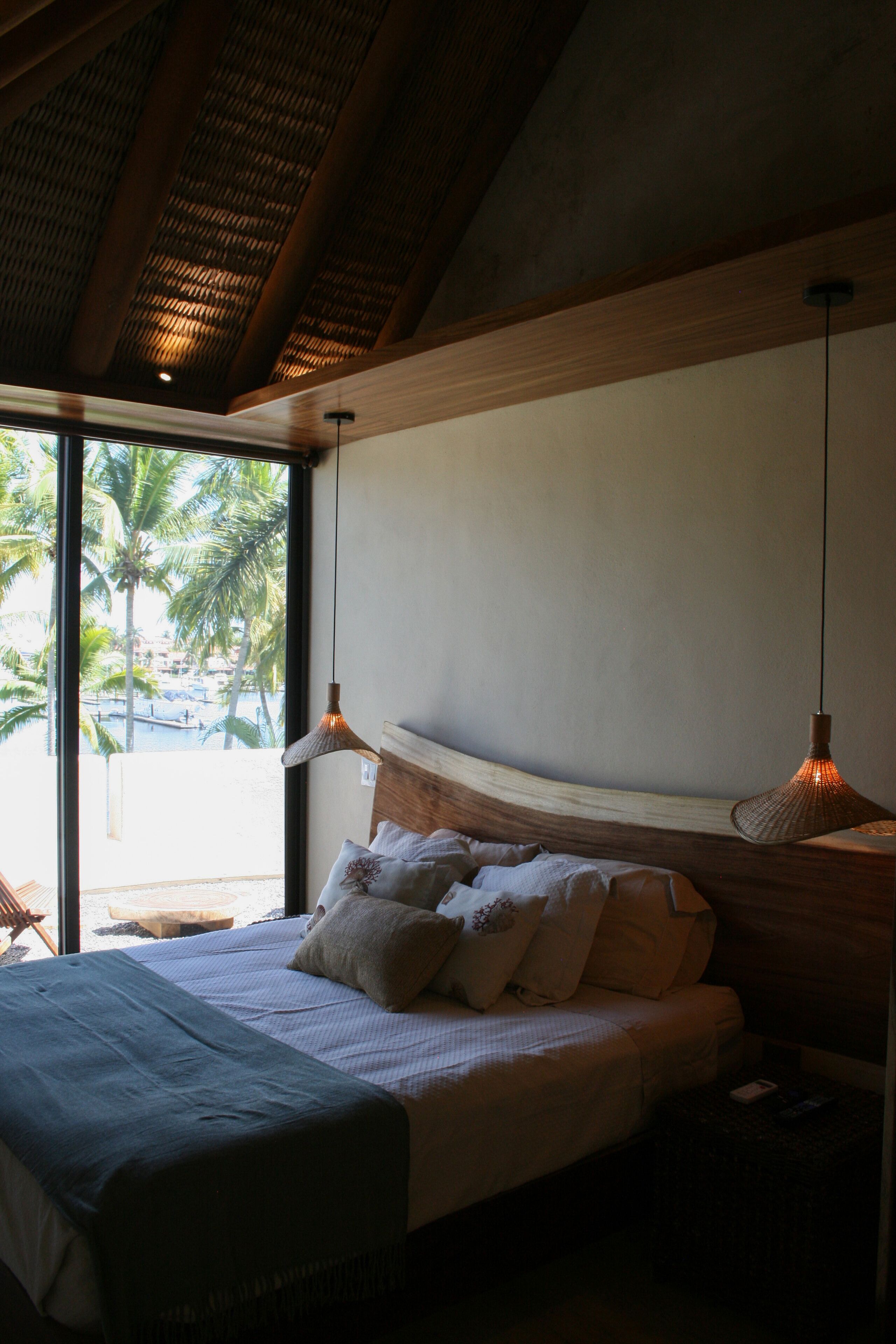 Une chambre avec un grand lit, deux lampes de chevet, une vue sur des palmiers et un plafond en bois.