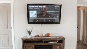 Un salon avec un téléviseur à écran plat fixé au mur, qui affiche la pochette d’un album de musique. Sous le téléviseur, on trouve un meuble de rangement en bois intégrant une cheminée.