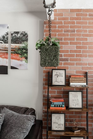 Un salon avec un mur en briques, une plante suspendue, une bibliothèque remplie de livres et de photos encadrées, et un canapé en cuir.
