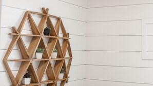 Un coin d’une pièce aux murs recouverts de panneaux blancs et au plafond en bois. Une étagère en bois aux formes géométriques est fixée au mur et supporte plusieurs petites plantes en pot.