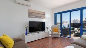 Ein Wohnzimmer mit Fernseher, Sofa und Sessel, einem großen Fenster mit Ausblick und einem Deckenventilator.