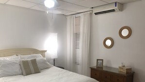 Un llit ben arropat amb llençols blancs, una taula de nit amb un marc de foto i un petit recipient, un aparador de fusta i un ventilador de sostre.