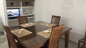 Una zona de menjador amb una taula i cadires de fusta, una televisió a una estant i una planta en test.