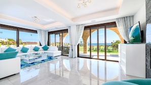 Un salon spacieux avec un canapé blanc, des coussins turquoise, une table basse en verre et un grand téléviseur posé sur un meuble en marbre.