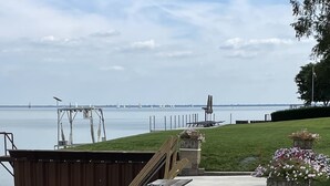 An outdoor patio with metal chairs and tables, a grassy area, and a view of a body of water with a bridge in the distance. - Lake Huron Beach  (Fort Gratiot Twp)