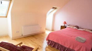 Une chambre avec un plafond en pente, un radiateur, un plancher en bois, un lit recouvert d’une couvre-lit rose, une table de chevet avec une lampe et une chaise avec un coussin rouge.