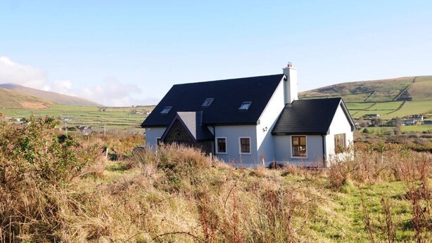 Une maison blanche avec un toit sombre, entourée de champs verdoyants et de collines.