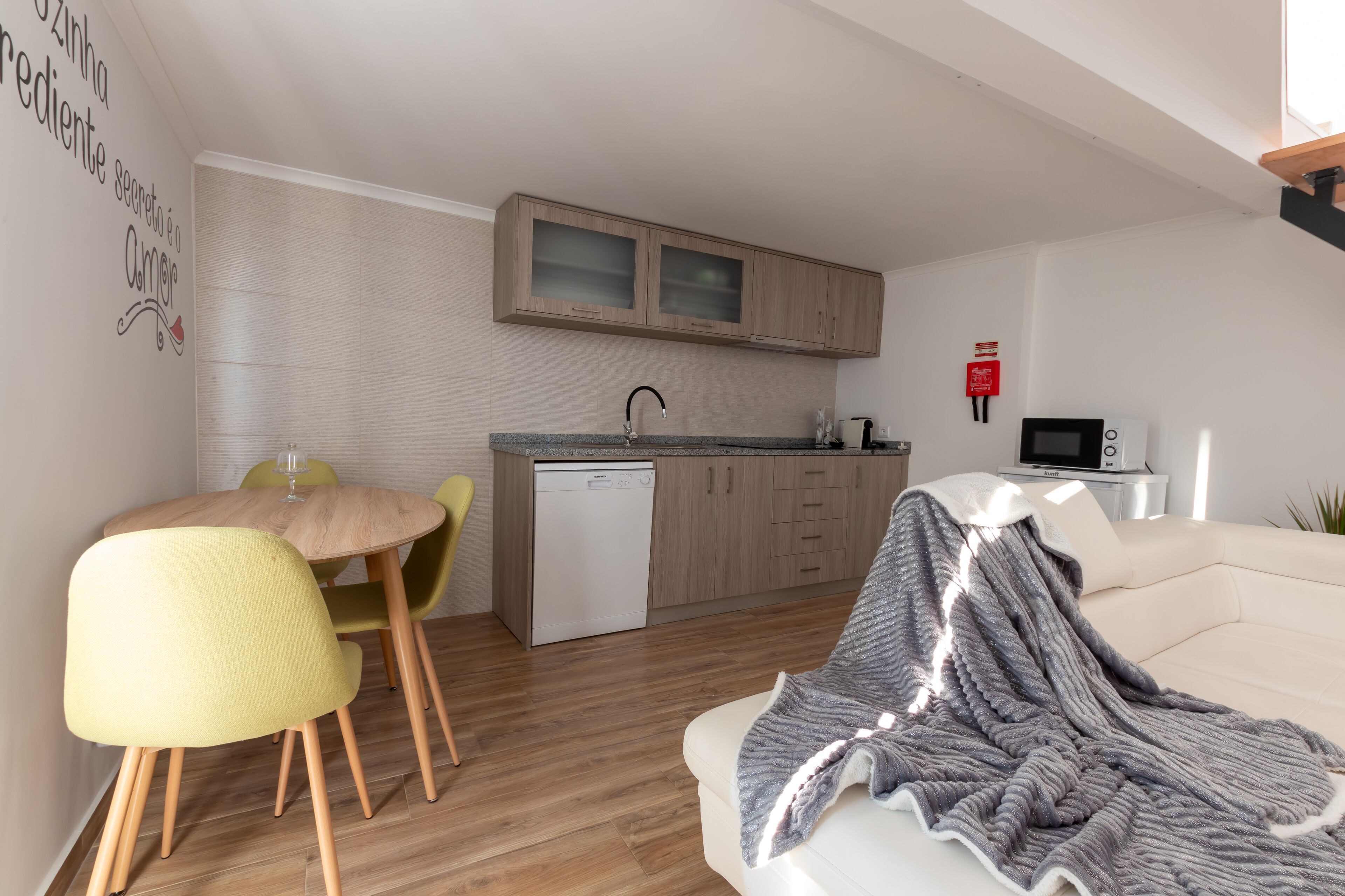 A modern kitchen with wooden cabinets and a dishwasher. There is a dining area with a round table and yellow chairs. A white sofa with a blanket is in the living room.