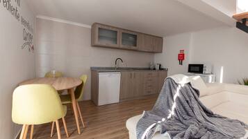 A modern kitchen with wooden cabinets and a dishwasher. There is a dining area with a round table and yellow chairs. A white sofa with a blanket is in the living room.