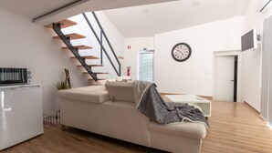 A modern living room with a white sofa, a wooden staircase, a kitchen area with a white refrigerator, and a wall clock. - Villa Alzira (Seia)