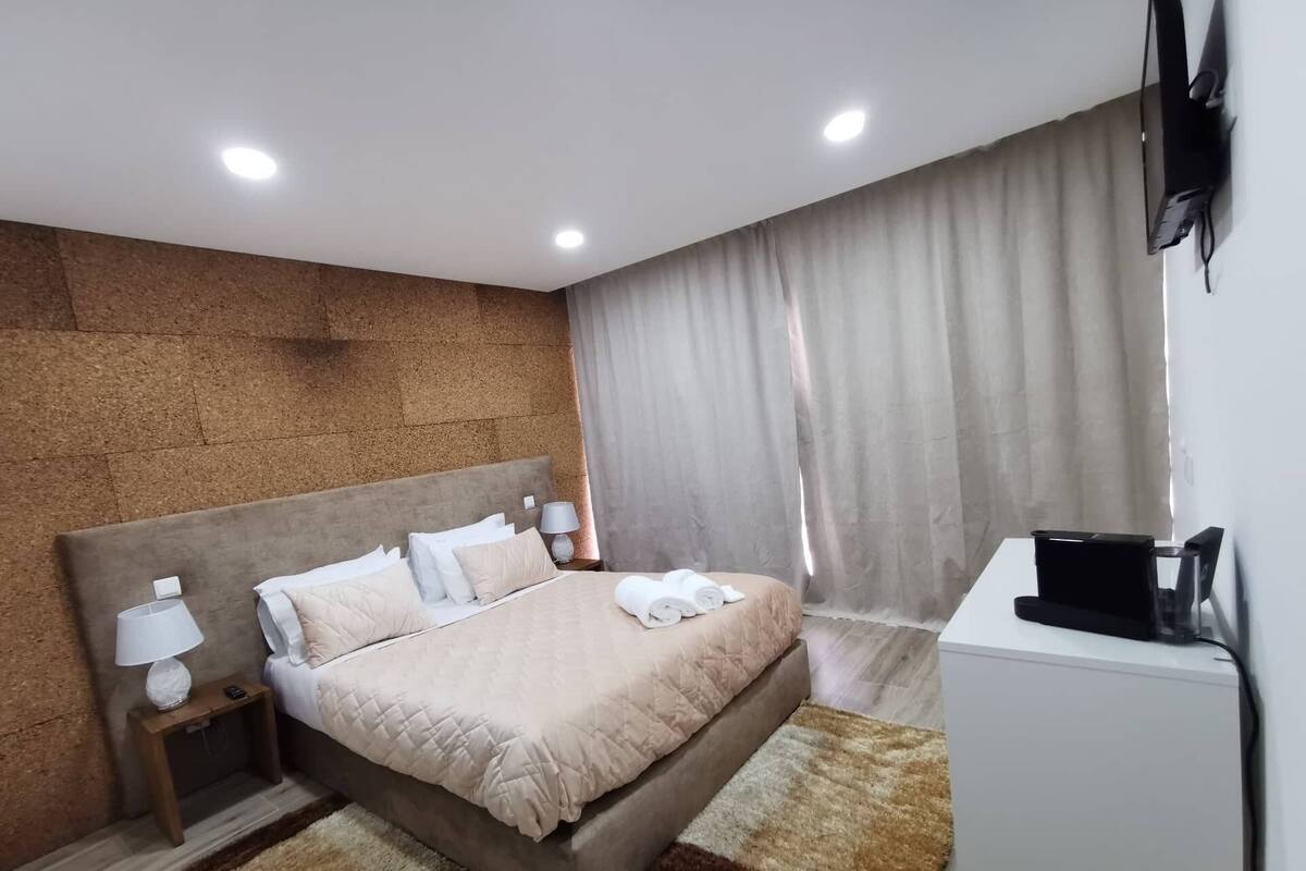 A modern hotel room with a large bed, a bedside table with a lamp, a small desk with a coffee machine, and a TV mounted on the wall.