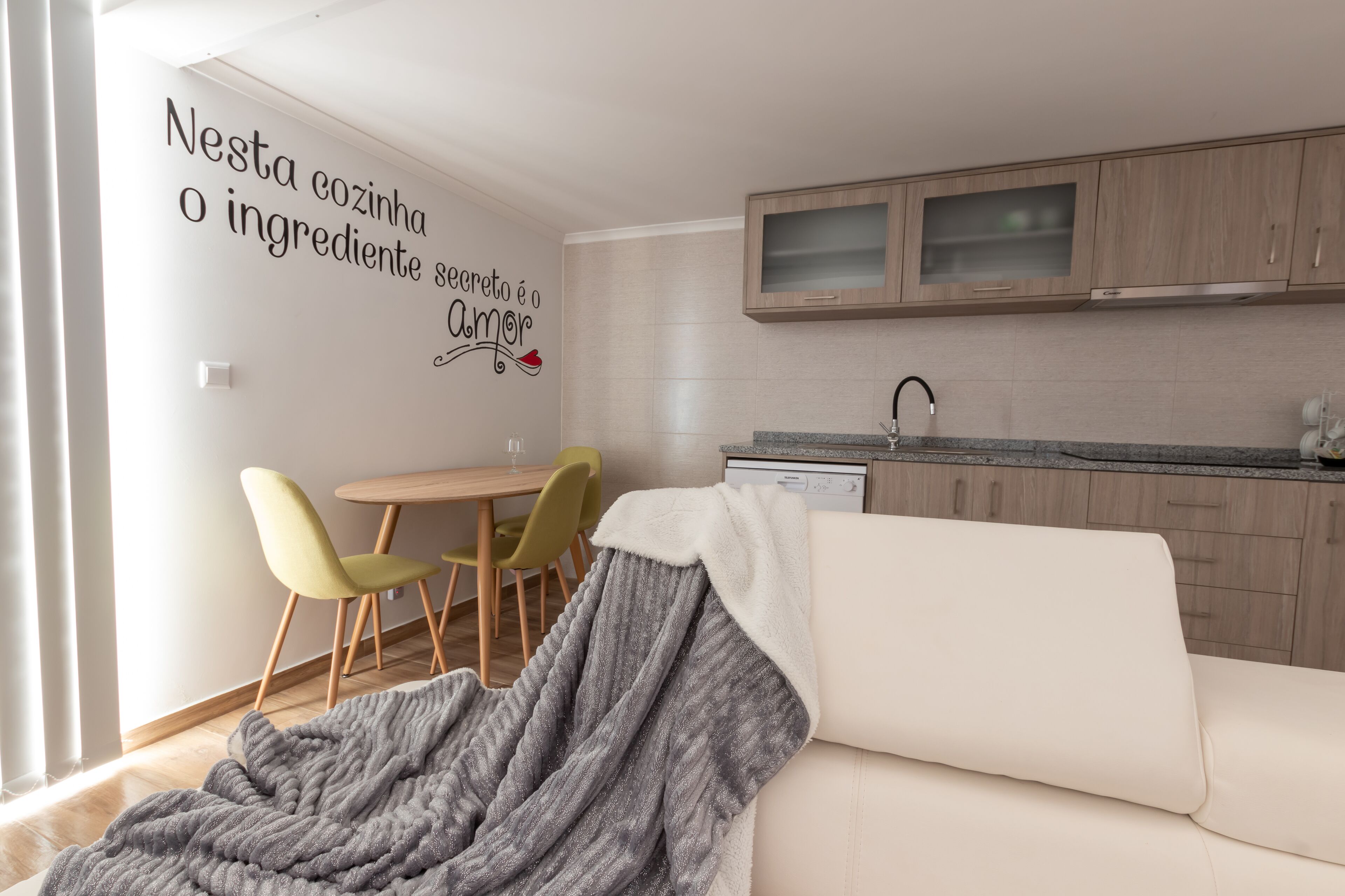A modern kitchen with wooden cabinets, a dining area with a table and chairs, and a cozy living space with a sofa and a blanket.