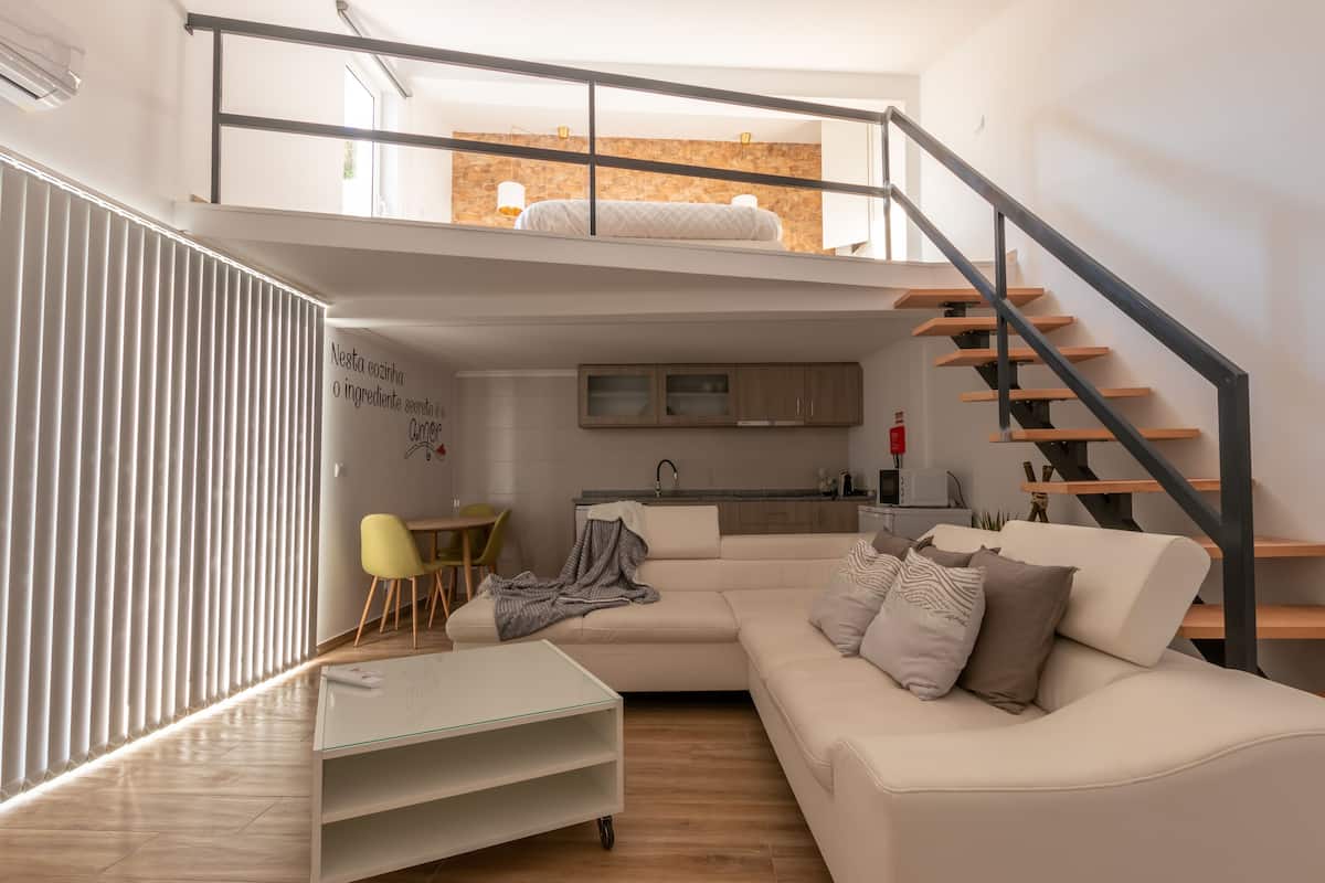 A modern living room with a sofa, a glass coffee table, a dining area with chairs, a kitchen with a sink, and a staircase.