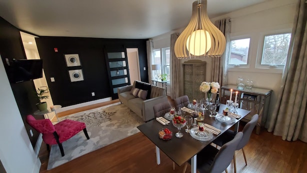 A modern living room with a dining area, featuring a wooden dining table set for a meal, a comfortable sofa, and a large window with curtains.