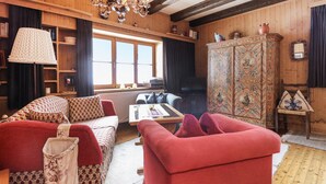 A cozy room with a wooden floor, a sofa with floral cushions, a red armchair, a cabinet with decorative panels, and a window with curtains. - barrierefreie Ferienwohnung (Sankt Veit/Defereggenta)