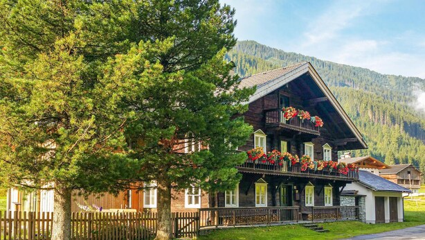 A traditional wooden chalet with a balcony adorned with flowers, set against a backdrop of a forested mountain. - barrierefreie Ferienwohnung (Sankt Veit/Defereggenta)