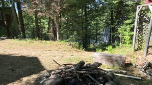 A campfire surrounded by rocks, a wooden bench, and a wooden structure in a forested area. - Waterfront Home for Summer/Winter Fun (Wakefield)