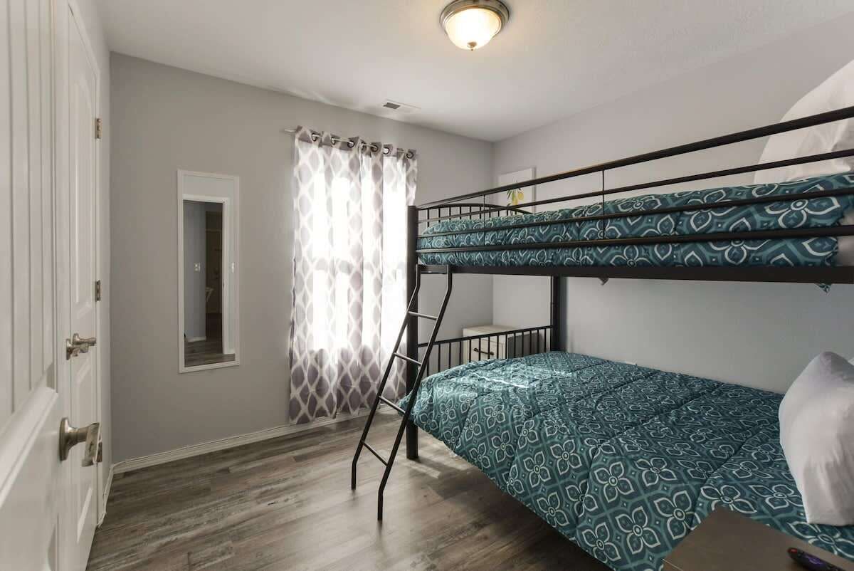 A bunk bed room with a patterned bedspread, a ladder, a window with curtains, and a door with a mirror.