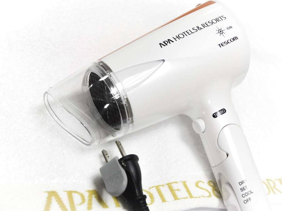 Un secador de pelo blanco con depósito de agua transparente, un enchufe y una toalla con el texto 'APA HOTELS & RESORTS'.